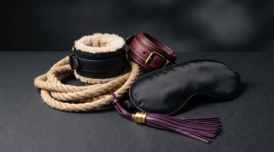 Composition élégante d'accessoires BDSM premium incluant menottes en cuir, corde de shibari, bandeau en satin et fouet, arrangés artistiquement sur fond noir.